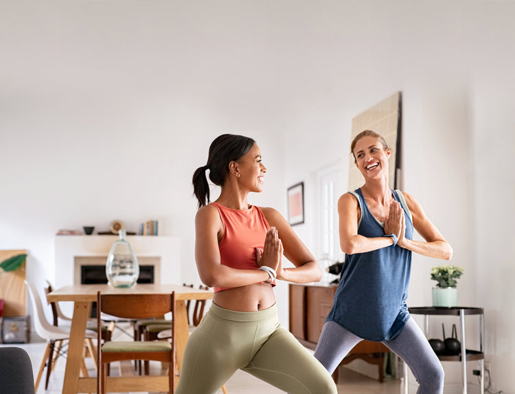 Two women in workout clothing, side by side in a lunging yoga pose, smiling at each other. A table and chairs sit behind them, along with various other household objects.