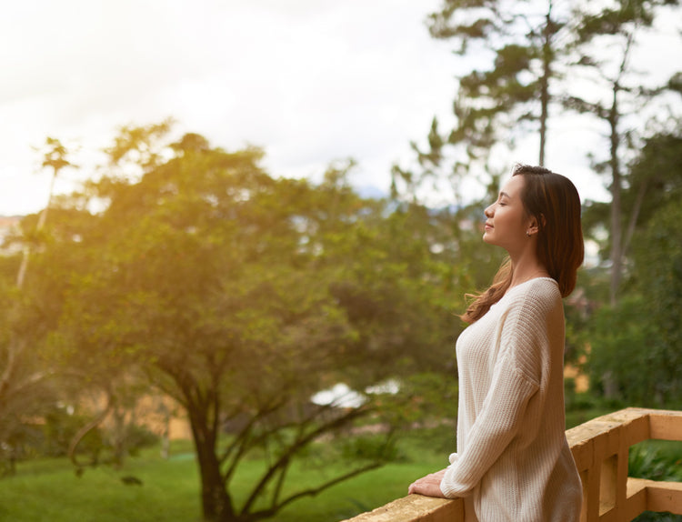 A woman standing outside on a wooden deck with her eyes closed, appearing to be taking a deep breath. There are many trees in the background, and a sun glare warms the image.