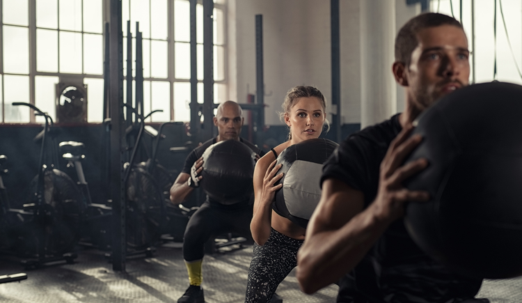 Three people lined up at the gym, each squatting and holding a medicine ball.