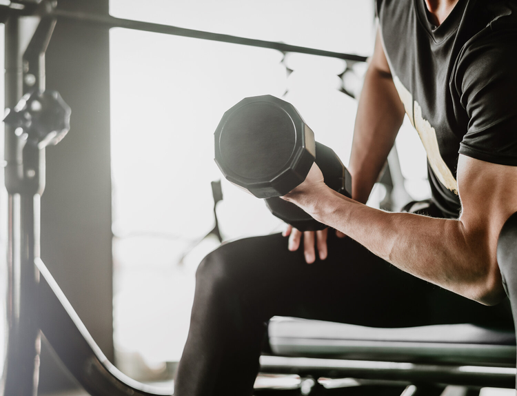 The muscular arm of someone sitting on a bench and doing a bicep curl with a dumbbell.