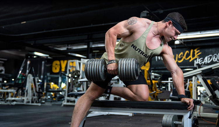 A man at the gym kneeling on a bench with one knee, lifting a large dumbbell with his opposite arm.