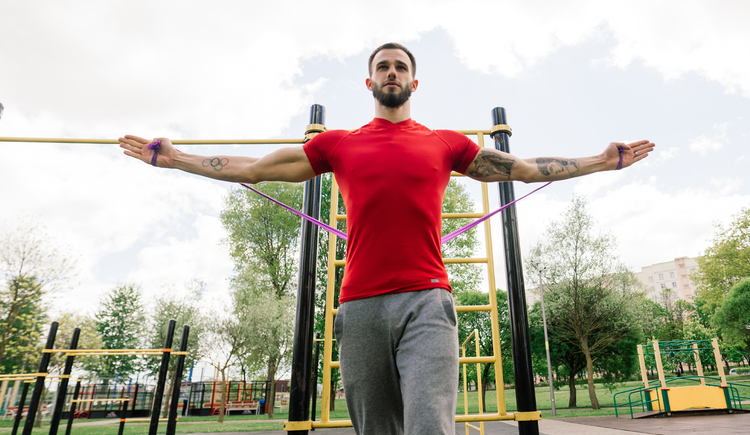 A man in a red T-shirt using resistance bands to exercise outside.