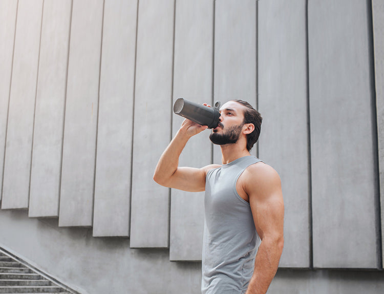 A man in a grey muscle tank, drinking from a black shaker bottle.