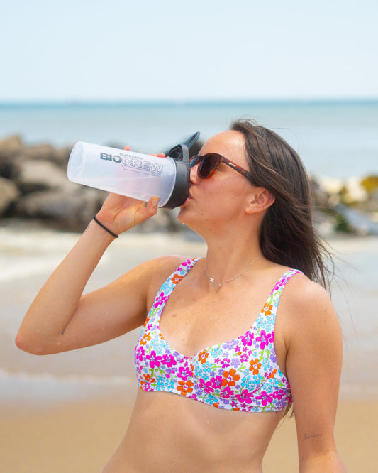 A woman in a floral bikini drinks from a "BioCrew" shaker bottle while standing on a beach near the ocean.