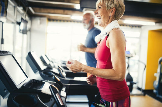 Older woman smiling while exercising on a treadmill in a bright fitness center.