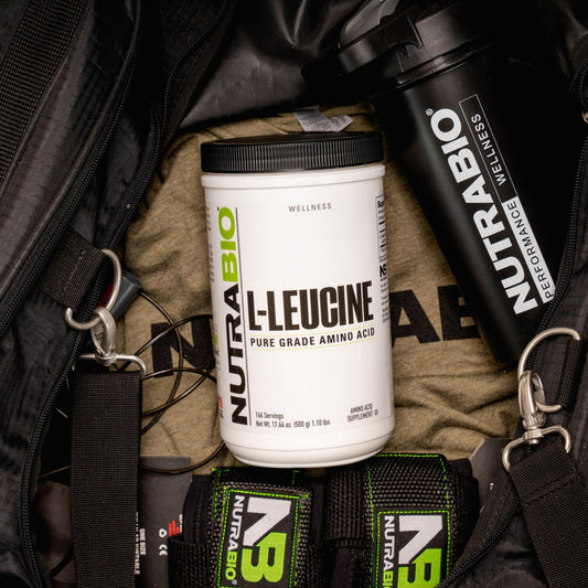 A white container of NutraBio L-Leucine powder sits inside a gym bag alongside a shaker bottle, lifting straps, and workout gear.