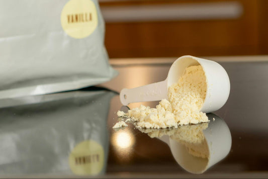 A white scoop of vanilla protein powder lies tipped over on a reflective surface beside a silver bag labeled "Vanilla."