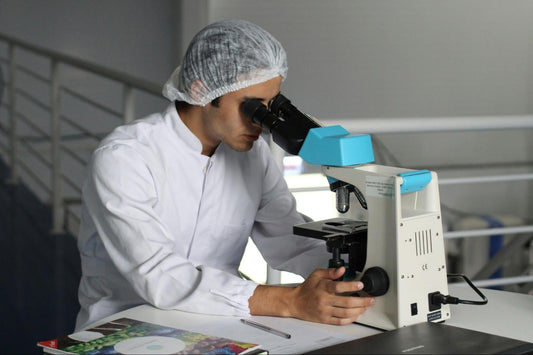 Scientist in a lab coat and hairnet looks through a microscope in a laboratory setting.