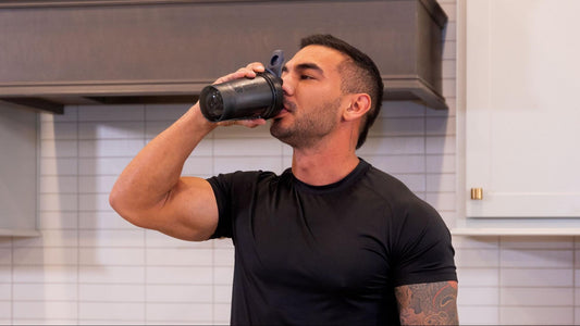 A muscular man in a black shirt drinks from a shaker bottle in a modern kitchen.