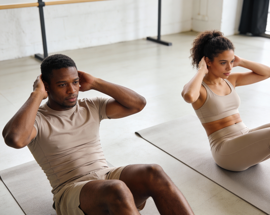 A man and woman in matching beige workout attire perform sit-ups side by side on yoga mats in a bright fitness studio.