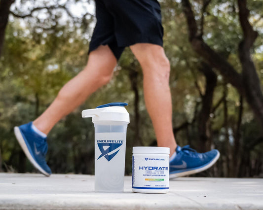A person walks outdoors in athletic gear with an EndurElite shaker bottle and a container of Hydrate Elite supplement placed on the ground in the foreground.