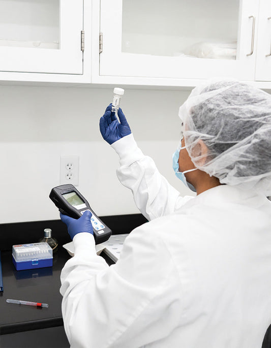 Lab technician in protective gear holds a small vial up to the light while operating a handheld measuring device at a laboratory bench.