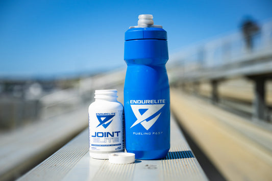 A blue EndurElite water bottle and an open bottle of EndurElite Joint Elite supplements sit on metal bleachers under a bright blue sky.