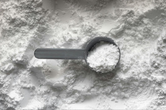 A gray plastic scoop is filled with fine white powder and placed on a surface covered in the same powder.