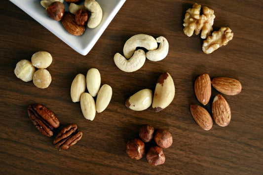 An assortment of nuts including almonds, cashews, walnuts, pecans, and macadamias is spread on a wooden surface with some in a white dish.