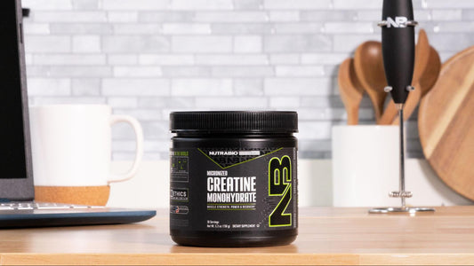 A container of NutraBio Creatine Monohydrate sits on a wooden kitchen counter, surrounded by a laptop, mug, and utensils in a clean, modern setting.