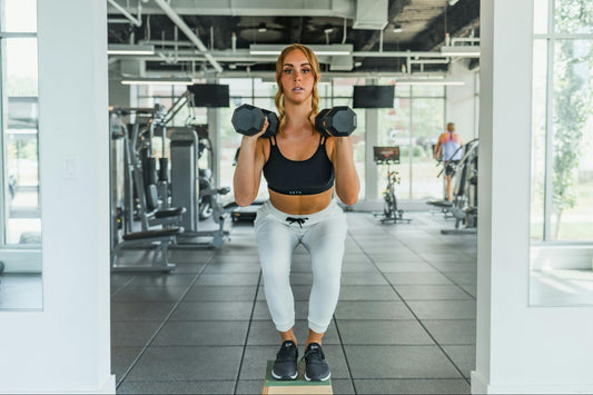 A woman in athletic wear performs a weighted step-up with dumbbells in a well-lit gym.