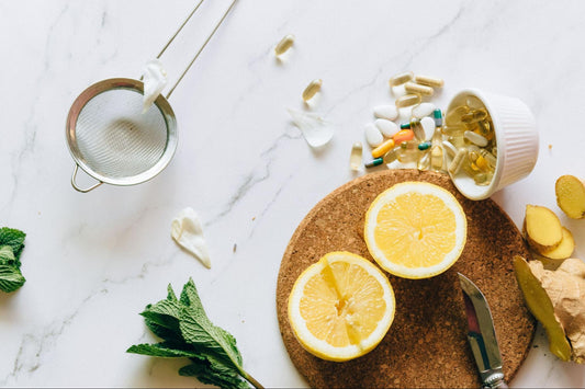 An overhead shot of two sliced lemon halves, fresh ginger root, and mint leaves on a cork mat next to a small cup spilling out various vitamin capsules and pills.