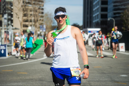 A male runner wearing a white "EndurElite" tank top proudly holds a finisher's medal after completing a race, with other runners and a cityscape in the background.
