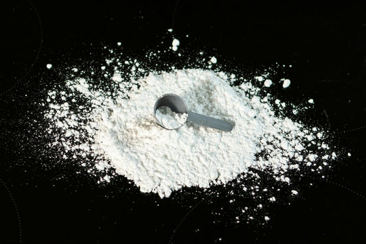 A gray plastic scoop rests on a scattered mound of white powder against a dark black surface.