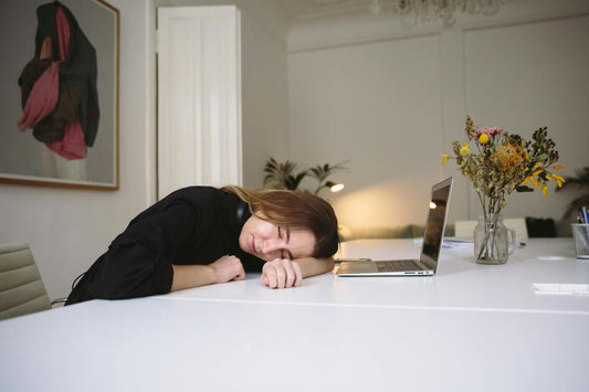 A woman rests her head on a table beside an open laptop, appearing tired in a softly lit room.