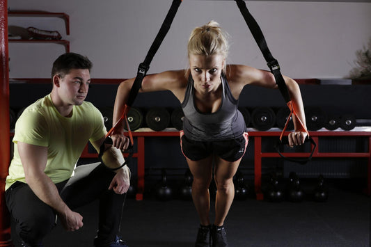 Woman training in the gym while a man squats beside her and watches