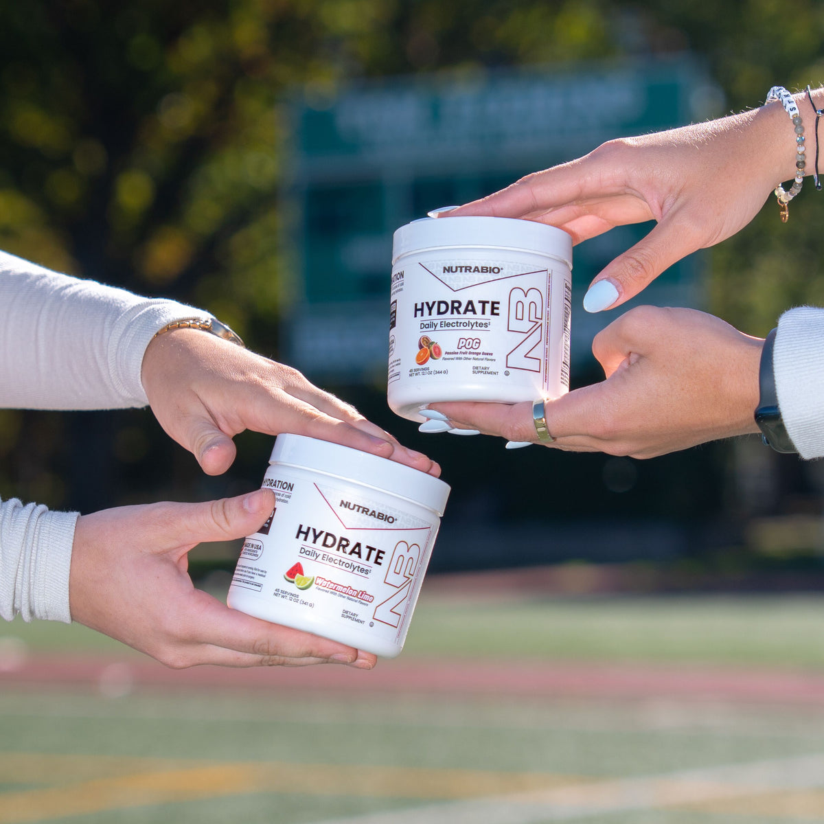 Two people holding white jars of Nutrabio Hydrate daily electrolyte powder in POG and Watermelon Lime flavors.