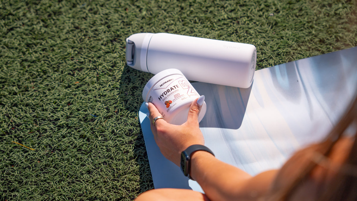 A hand holding a tub of Nutrabio Hydrate electrolytes next to a white water bottle on a yoga mat.