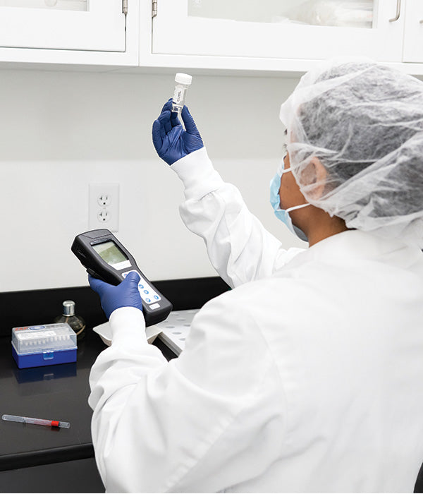 A scientist in protective clothing, gloves, and a hairnet holds a vial in one hand and a handheld device in the other while working in a laboratory. Lab equipment and supplies are on the counter nearby.