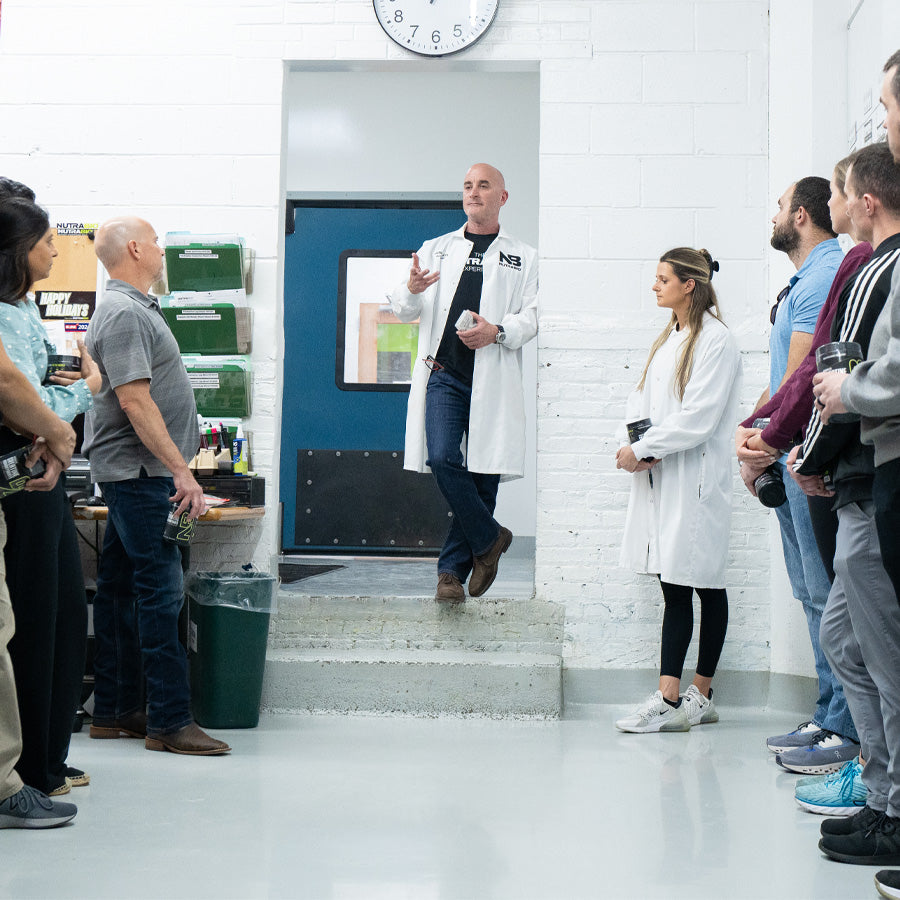 A man in a lab coat speaks to a group of people gathered in a bright, industrial room. Another person in a lab coat stands nearby, while others listen attentively. A clock hangs on the wall above the speaker.