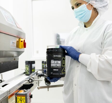 A person in a lab coat, hair net, face mask, and gloves holds a container labeled Creatine Monohydrate near industrial equipment in a clean, sterile laboratory setting.